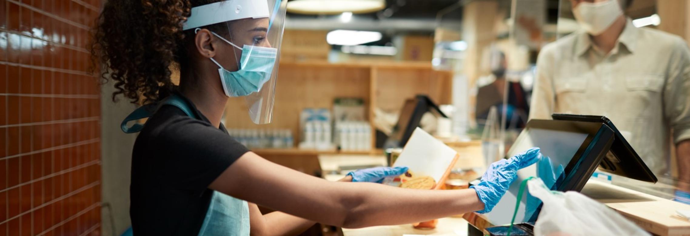 a cashier in protective gear rings up a customer at a grocery store